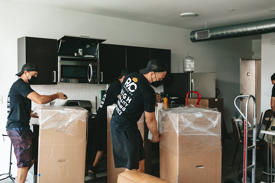 A close-up of a person wearing a grey hoodie assembling furniture, using a screwdriver to attach metal brackets to a wooden surface. The workspace is indoors, with a cardboard box and packing material