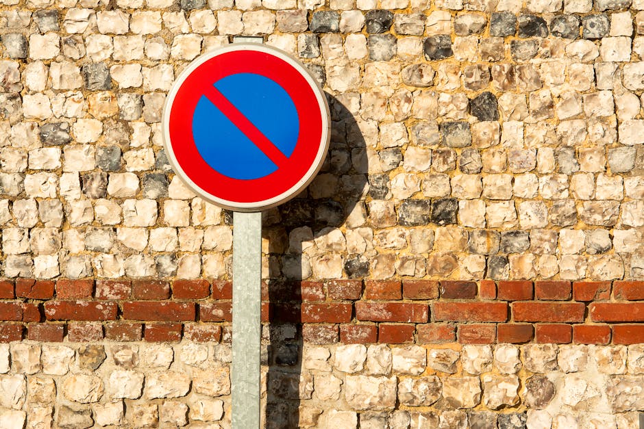 A close-up view of a rectangular white and red 'No Parking' sign mounted on a metal pole, which is situated behind a concrete curb. The sign features a black letter 'P' with a red circle and diagonal
