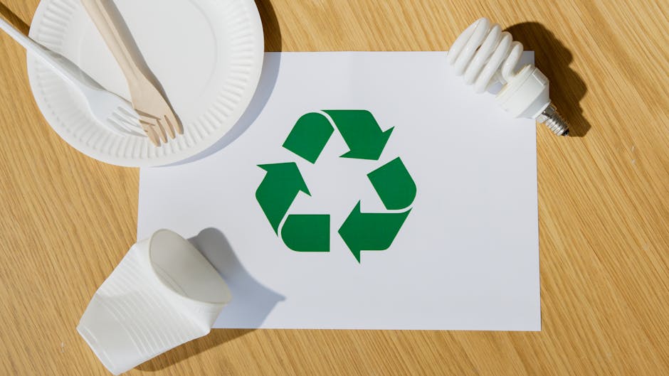 A photograph showing a wooden surface with various household items arranged around a white sheet of paper featuring a green recycling symbol. On the left side, there is a white paper plate holding woo